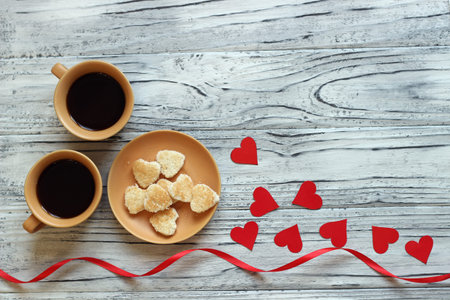 On a wooden background there are two cups of coffee next to a saucer with cookies in the form of...