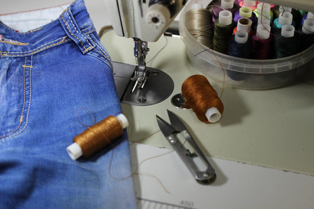 On the table of the sewing machine lie denim fabrics, close-up, next to the thread and scissors.
