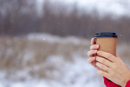 A young woman in a red jacket in winter holds a glass of hot coffee or tea. A snowy winter and a hot...