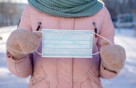 A girl in a jacket and hat on the street in winter holds a protective medical mask during the coronovirus pandemic.の写真素材