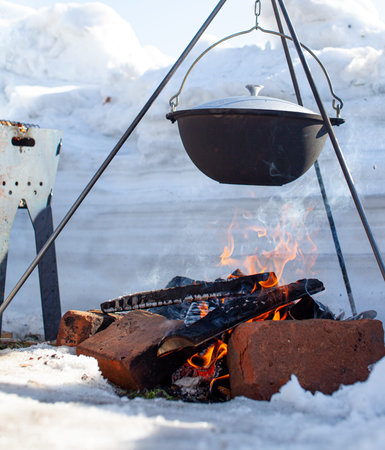 Over the fire hangs a pot in which to cook food. On a hook on a tripod, steam comes out of the pan....