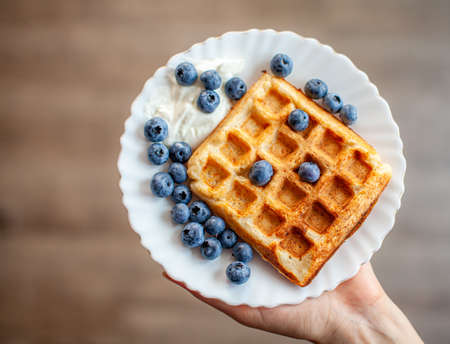 A woman holds in her hand a plate on which fresh waffles with berries are ready. Homemade waffles with strawberries and blueberries. Delicious Breakfast with berries.の写真素材