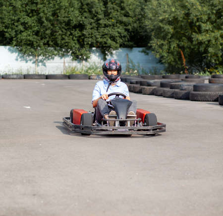 A girl or a woman in a hard hat rides a go-kart on a special track fenced with rubber wheels. Active recreation and sports on transport. Preparation and training for competitions.の写真素材