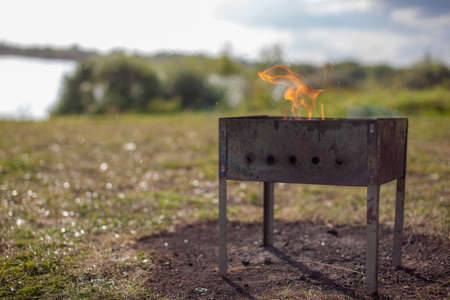 The firewood in the grill burns with a bright orange flame of fire on a natural green background. Preparation for cooking meat on the grill in nature. Fire flames and smokeの写真素材