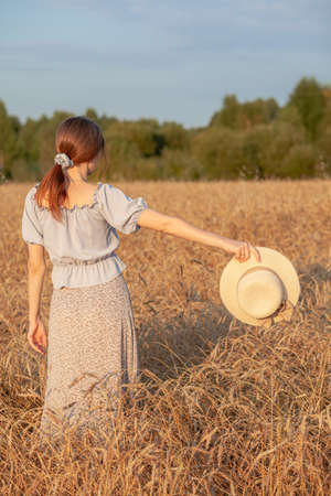 Young beautiful girl with long curly hair poses in a wheat field in the summer at sunset. A girl holds a hat in her hand against the background of a wheat field. The view from the back. Toning.の写真素材