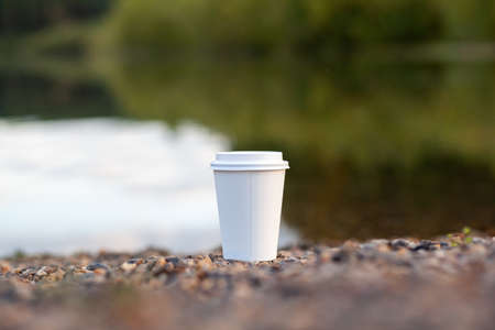 A white paper cup of coffee or tea stands on the rocky shore of the lake. A mug of hot drink on a pebble. Beautiful nature background of the lake in the forest behind. free copy space.の写真素材