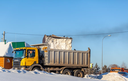December 23, 2021. Kemerovo Region, Russia. Big orange tractor cleans up snow from the road and loads it into the truck. Cleaning and cleaning of roads in the city from snow in winter.のeditorial素材