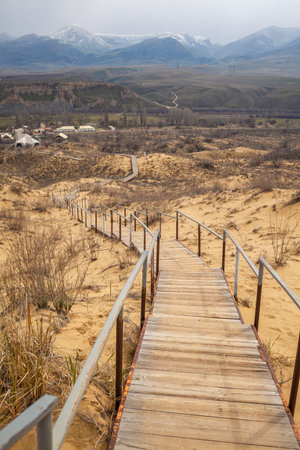 The staircase rises to a sandy dune. Climbing to the top in the desert. Sarykum dune. Dagestan, Russia. A unique sandy mountain in the Caucasus on a cloudy day. Grass grows on a sand dune.の写真素材