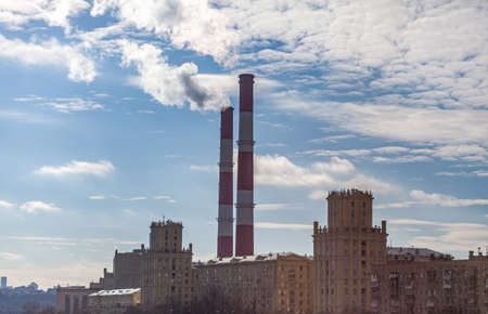 CHP in the city. Smoke rises into the sky from the heat pipes of the power plant. Pipe of combined heat and power plant CHP or cogeneration against background of multi-storey residential buildingsの写真素材