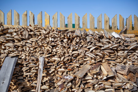 Firewood is stacked in a woodpile along the fence against the blue sky