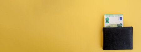 A hundred euro banknote sticks out of a black wallet on a yellow background. cash paper currency,...