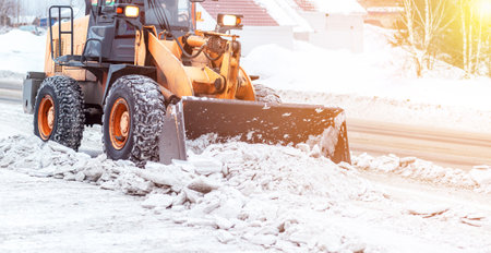 Orange tractor cleans up snow from the road and loads it into the truck. Cleaning and cleaning of roads in the city from snow in winter. Snow removal after snowfall and blizzards.の写真素材