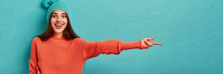 A woman wearing a knit hat and sweater excitedly points to an empty space on a turquoise background. She appears to be advertising or promoting something.の素材