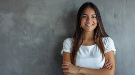 A portrait of a young Hispanic woman with a pleasant smile and crossed arms, standing against a gray wall.の素材