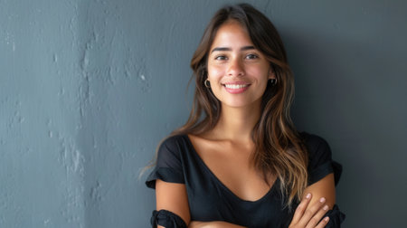 A young Latina woman with long brown hair smiles cheerfully at the camera. She stands against a gray wall with her arms crossed, creating a confident and approachable vibe.の素材