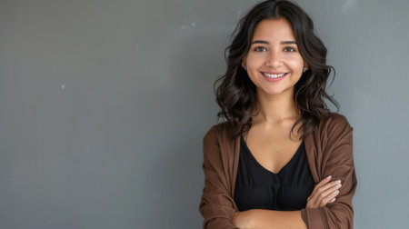 Portrait of a young Hispanic woman with a pleasant smile, standing with crossed arms against a gray wall. She is looking directly at the camera.の素材