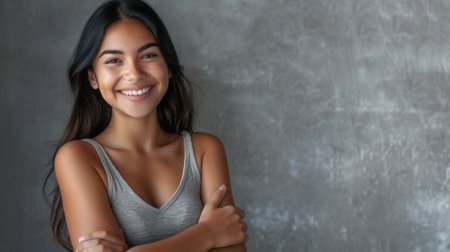 A portrait of a young Hispanic woman smiling brightly while standing against a gray wall with her arms crossed.の素材