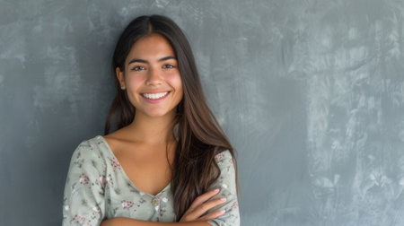 A young Hispanic woman with long dark hair smiles confidently at the camera while standing against a gray wall with her arms crossed.の素材