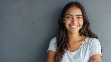 A young Hispanic woman with long dark hair smiles cheerfully at the camera while standing with her arms crossed against a gray wall.の素材