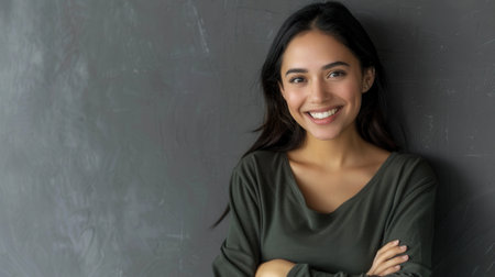 A portrait of a young Hispanic woman with long black hair and a warm smile. She is standing against a gray wall with her arms crossed, looking directly at the camera.の素材