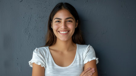 A young Hispanic woman stands against a gray wall with a pleasant smile. She has her arms crossed and is looking directly at the camera.の素材