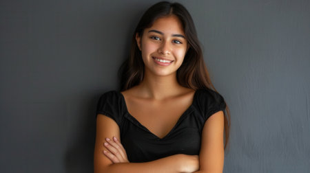 A young Hispanic woman with long dark hair smiles brightly while standing against a gray wall with her arms crossed.の素材