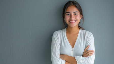 A portrait of a young Hispanic woman with a pleasant smile, standing with her arms crossed against a gray wall.の素材