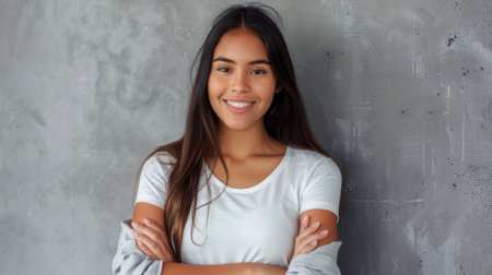 A young Latina woman smiles cheerfully at the camera with her arms crossed. She stands against a gray concrete wall with copy space.の素材