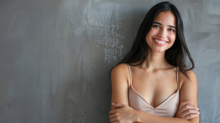 A young Latina woman stands with her arms crossed, smiling brightly while looking directly at the camera. She is positioned against a gray wall with copy space.の素材