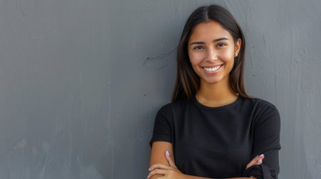 A young Hispanic woman with long brown hair smiles brightly while standing against a gray wall with her arms crossed.の素材