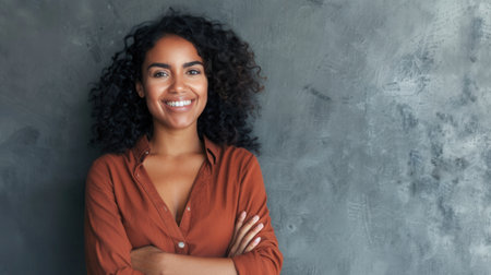 A young Hispanic woman with curly hair and a pleasant smile stands against a gray wall with her arms crossed.の素材