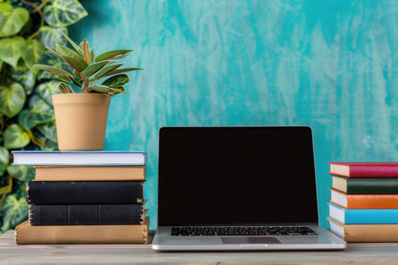 A laptop computer and a stack of textbooks sit on a desk in front of a teal wall, creating a contemporary image of modern learning.の素材