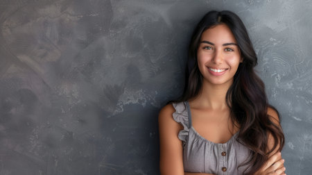 A young Hispanic woman with long black hair smiles cheerfully at the camera while standing against a gray wall with her arms crossed.の素材