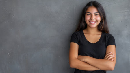 A young Hispanic woman with long, dark hair stands against a gray wall, smiling cheerfully with her arms crossed.の素材