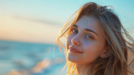 A close-up portrait of a young woman with long blonde hair smiling at the camera while standing at the beach on a sunny summer day.の素材