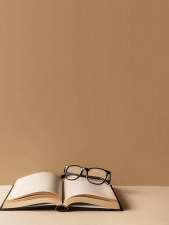 A book lies open on a table, with a pair of black glasses resting on the pages. The background is a solid brown color.の素材