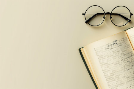 A pair of black, round glasses lie atop an open book on a white table.の素材
