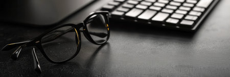 A pair of black eyeglasses sits on a dark desk beside a computer keyboard.の素材