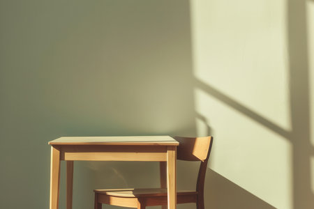 A wooden table and chair sit near a wall, sunlight streaming through a window casts shadows on the wall.の素材