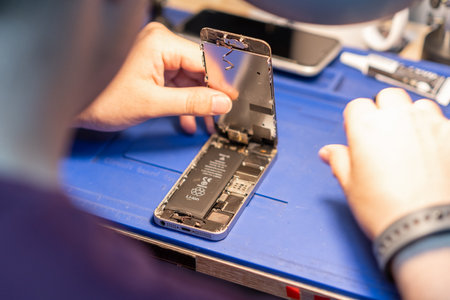 A technician is meticulously replacing the battery of a smartphone in a well-organized repair workshop filled with tools.の写真素材