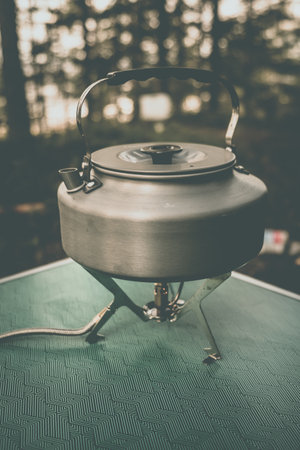 A silver kettle is placed on a camping stove surrounded by trees, showcasing a serene outdoor cooking setup.の写真素材