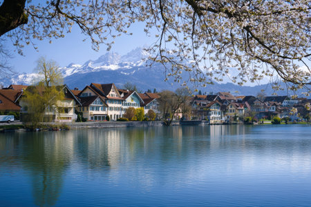 Cherry blossoms frame charming houses reflecting in the serene lake, with majestic snow-capped peaks visible in the distance.の素材