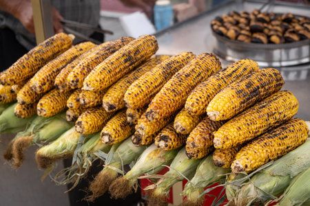 A variety of grilled corn on the cob piled high for sale at a bustling summer market, showing bright...