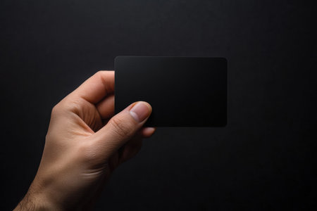 A hand elegantly displays a sleek black gift card, set against a dark backdrop, offering space for personalized text and design elements.の素材