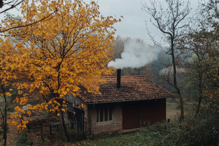 A secluded cabin nestled among vibrant autumn foliage with smoke gently ascending from the chimney, showing the peaceful countryside setting.の素材