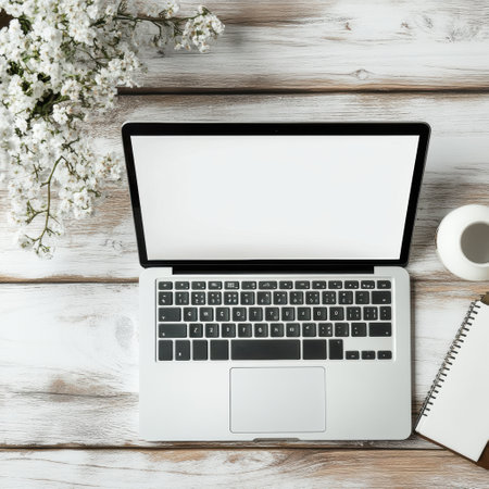 A laptop rests on a wooden table alongside a notebook and a coffee cup, with fresh flowers adding a touch of nature to the workspace.の素材
