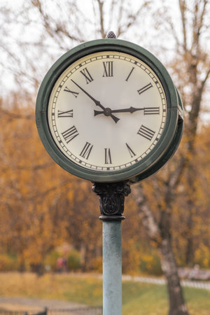 A vintage clock mounted on a pole displays the time amidst a backdrop of colorful autumn leaves,...