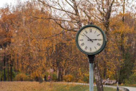 A vintage clock stands tall in a park, framed by vibrant autumn leaves as people stroll by in the...