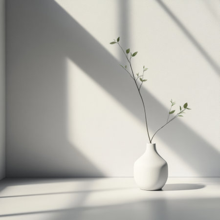 A simple white vase holds slender branches, illuminated by morning light, creating soft shadows on the wall in a stylish room.の素材