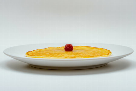 A thin golden pancake is placed on a white plate, topped with a single fresh raspberry, showing a simple yet appealing dessert.の素材
